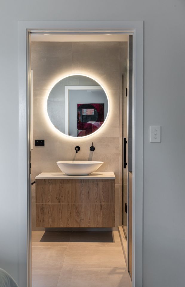 A compact contemporary bathroom viewed through an open doorway, featuring a floating timber vanity with a white vessel basin, black wall-mounted tapware, large beige stone-look wall and floor tiles, and a round backlit mirror. The bathroom is framed by a light grey painted wall and a glazed or reflective door panel.