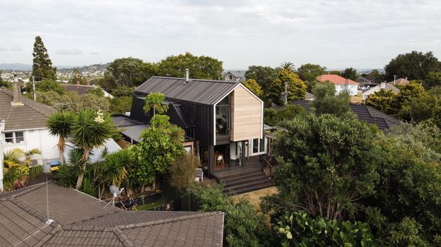 Elevated exterior view of a compact contemporary residential house set among established suburban gardens. The home has a steep gabled form, dark corrugated metal roof, warm horizontal timber-clad front gable, extensive glazing, and a timber deck and stair leading into the landscaped site. Neighbouring houses and mature vegetation surround the property.