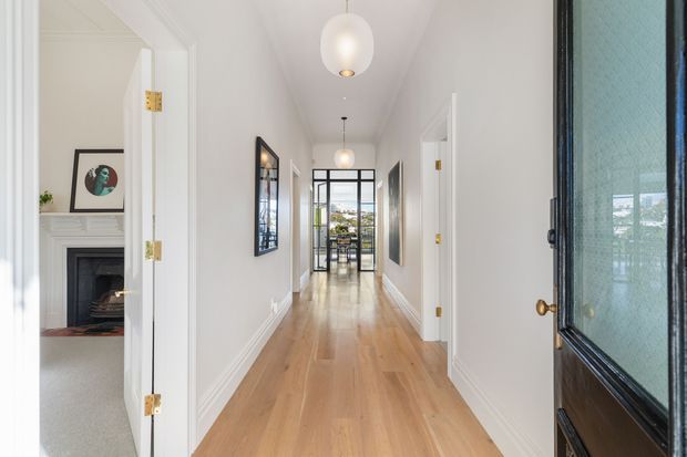 Bright, refined residential hallway viewed from an open front door, with wide pale timber flooring, white painted walls and traditional moulded skirting and architraves. The corridor leads to a glazed exterior door or window at the far end, with pendant lights overhead. A room with a classic fireplace and framed artwork is visible through an open doorway on the left, while additional white-trimmed doorways line the hall.
