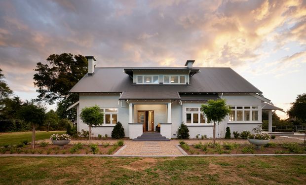 Front exterior view of a large, symmetrical gabled-roof home with white rendered walls, dark grey roofing, dormer windows, a covered entry porch, and formal landscaped gardens. The house is photographed at sunset, creating a warm, refined residential presentation.
