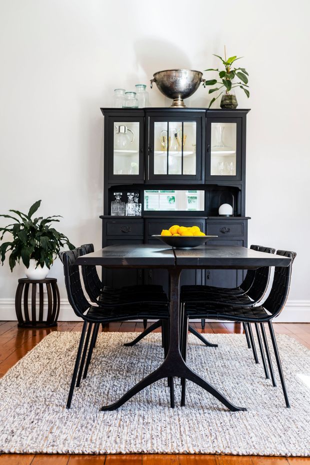 A bright, refined dining room featuring a dark wood or painted black hutch with glazed display cabinets, a dark dining table, and black woven dining chairs. A neutral textured rug sits over timber flooring, while plants, glassware, and a bowl of citrus add natural and colourful accents. The room has white walls and classic trim, with a balanced, high-contrast palette.
