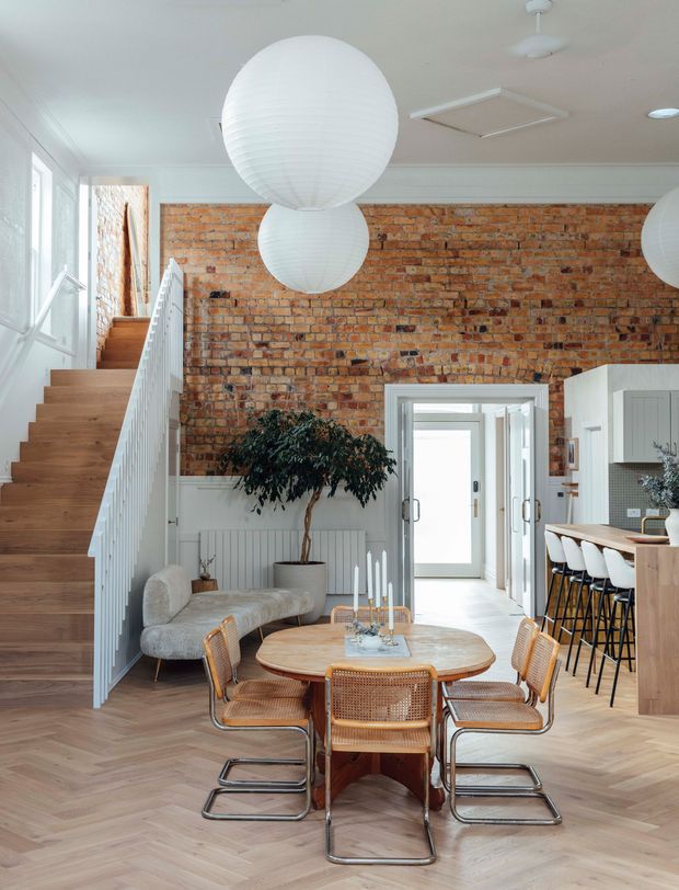 Bright open-plan dining and kitchen area with a prominent exposed red-brick feature wall, pale timber herringbone flooring, white cabinetry, and a timber dining table surrounded by woven cane chairs. A staircase with white balustrading rises along the left side, while large white paper pendant lights hang from the ceiling. A small lounge nook and indoor tree sit beneath the stair, with a hallway visible beyond.