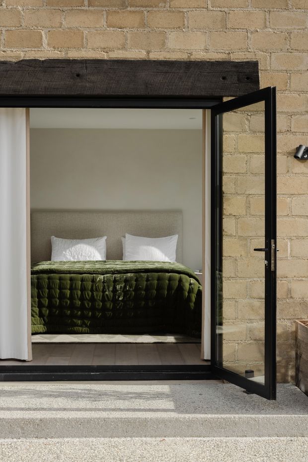 View from an exterior concrete step into a bedroom through an open black-framed glazed door. The room contains a bed with a tall upholstered beige headboard, white pillows, and a deep olive-green quilt. The surrounding building fabric features pale exposed brick, dark timber above the opening, black metal door framing, and a pale polished concrete threshold and steps. Strong natural sunlight creates a calm, refined indoor-outdoor composition.