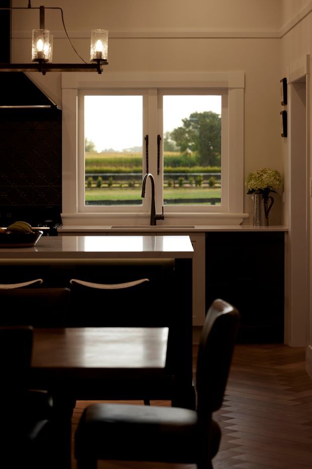 A warm, traditional kitchen with a central island, dark cabinetry, a pale stone benchtop, integrated sink and curved tap, black tiled splashback, and framed white double windows overlooking a rural landscape. A dining table and upholstered chairs occupy the foreground, with pendant lights overhead and timber parquet flooring.