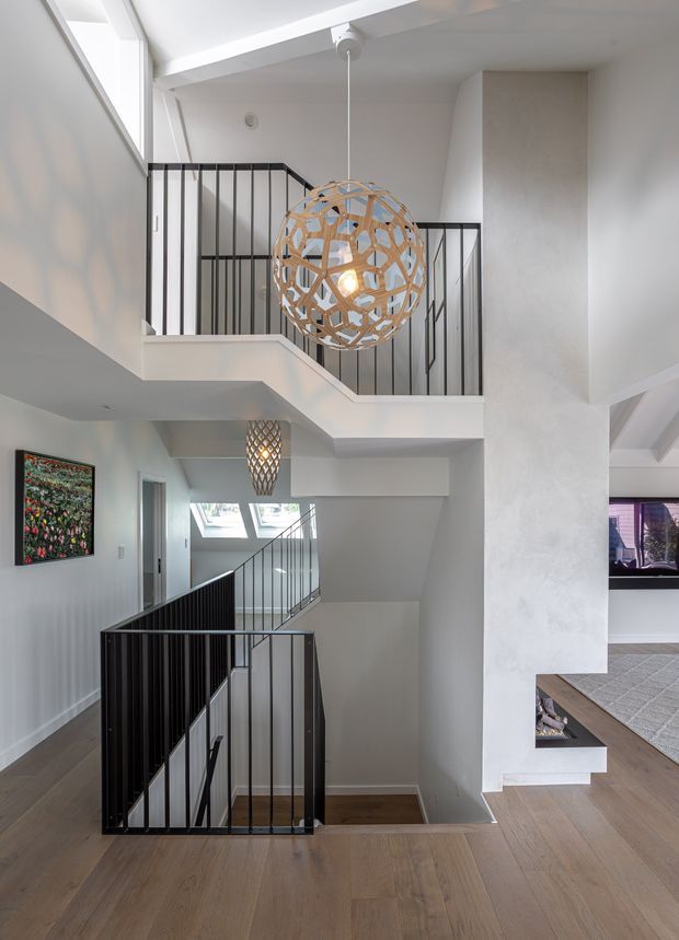 Bright contemporary multi-level stair landing and hallway with a stairwell enclosed by slim black metal balustrades. White walls and structural beams contrast with warm timber flooring, while large sculptural pendant lights hang through the stair void. Skylights and upper-level glazing bring in abundant natural light, and a living area is partially visible beyond the landing.