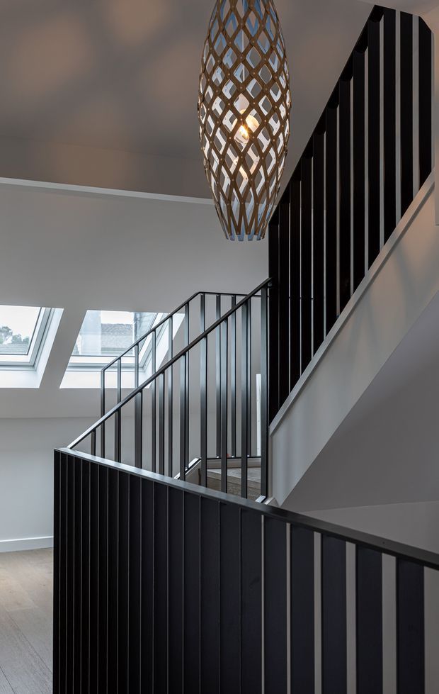 Interior architectural photograph of a contemporary stairway and landing with black vertical metal balustrades, pale walls and ceiling, skylights, and a sculptural woven pendant light. The staircase rises through a bright multi-level circulation space, with timber-toned flooring visible below.