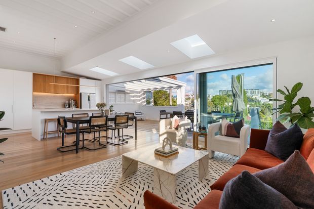Bright open-plan apartment interior combining a contemporary kitchen, dining area and living room. The space features pale timber flooring, white cabinetry, a timber kitchen accent wall, black-framed glazing, skylights, a marble-look coffee table and a large patterned rug. Floor-to-ceiling sliding glass doors open onto a covered terrace or balcony with an urban outlook. A rust-orange sofa and greenery add warmth and contrast.