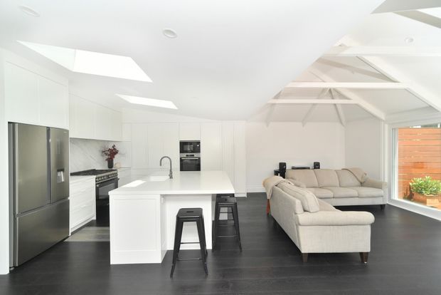 Bright open-plan kitchen and living area with white cabinetry, a large central island, stainless-steel appliances, dark timber flooring, and a light sectional sofa. Exposed white roof trusses and multiple skylights create an airy, high-ceilinged interior, with large glazing opening toward a timber privacy screen.