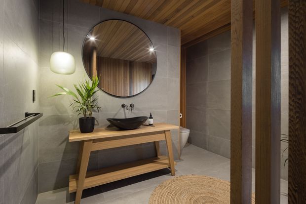 A warm, contemporary bathroom featuring large grey wall and floor tiles, a timber ceiling, timber-framed detailing, and a freestanding timber vanity console with a black vessel basin. A round mirror, pendant light, indoor plant, toilet, and walk-in shower area are visible. The space has a refined natural-material palette and layered ambient lighting.