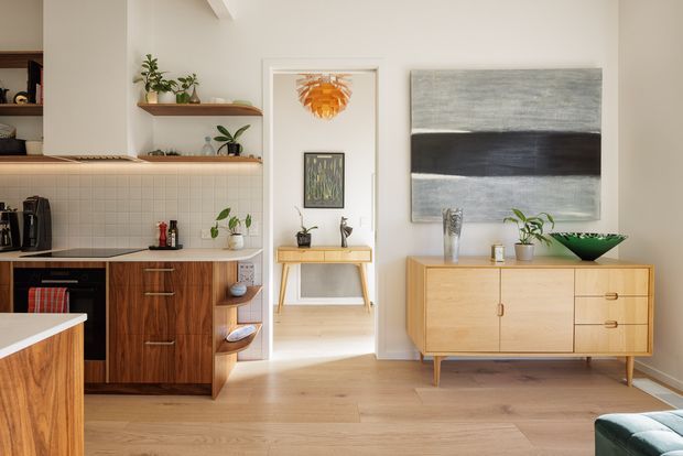 Warm, light-filled open-plan interior featuring a compact kitchen with dark timber cabinetry, pale tiled splashback, open shelving and a peninsula, connected to a living area with a mid-century-style timber sideboard, artwork and plants. Pale timber flooring and white walls create a calm, balanced setting.
