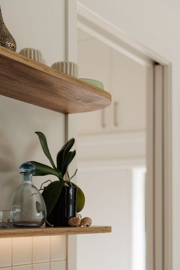Close-up interior detail of warm timber floating shelves displaying ceramics, a glass bottle, a potted plant and small decorative objects. The shelves are mounted against a light tiled wall beside a white-trimmed doorway, with a softly lit white kitchen or utility area visible beyond.