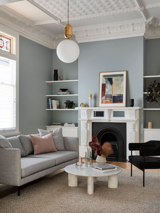 A refined living room with a grey upholstered sofa, black accent chair, round pale stone coffee table, and a decorative white fireplace with a black cast-iron insert. Built-in open shelving flanks the fireplace, while a large abstract artwork hangs above it. The room features muted blue-grey walls, ornate white ceiling cornices and ceiling roses, detailed fireplace mouldings, a pendant globe light with brass fitting, and tall windows with stained-glass transom detailing. The styling is elegant, calm, and contemporary within a period architectural shell.