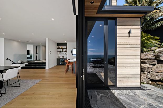Split architectural view showing a bright contemporary open-plan interior opening directly onto a landscaped exterior terrace. Inside are pale timber flooring, white walls and ceiling with recessed downlights, a dining area, open shelving, and a stair with dark detailing. Outside, a black-framed glazed door and clerestory-style glazing sit beside horizontal light timber cladding, with dark irregular stone paving and a rocky landscaped garden.