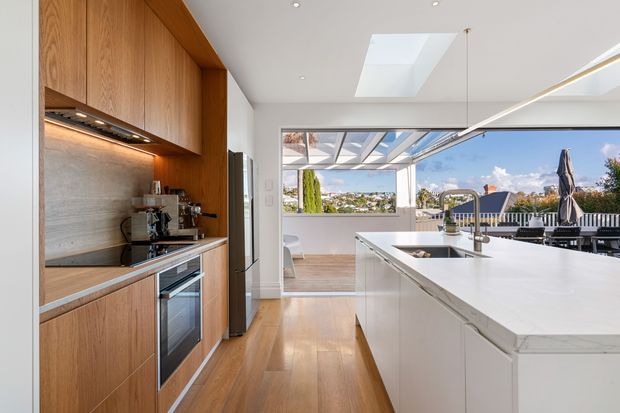 Bright contemporary kitchen with a long white island and integrated sink, warm timber cabinetry, stone benchtops, stainless-steel appliances, and a textured stone backsplash. The space opens to a glazed dining or outdoor entertaining area with views beyond, and includes a skylight, concealed under-cabinet lighting, and linear pendant lighting.