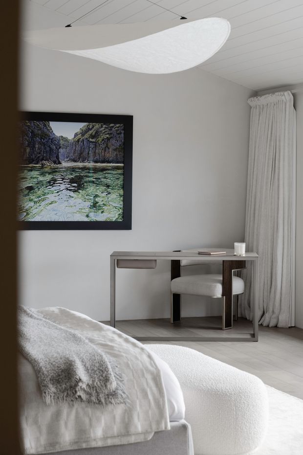 A softly lit, refined bedroom interior in a restrained neutral palette. The room features a low upholstered bed in the foreground with a textured white throw, a minimalist metal-framed writing desk and upholstered chair, full-height gathered curtains, pale walls and flooring, and a large framed coastal landscape artwork. A sculptural white pendant light hangs from a linear paneled ceiling.