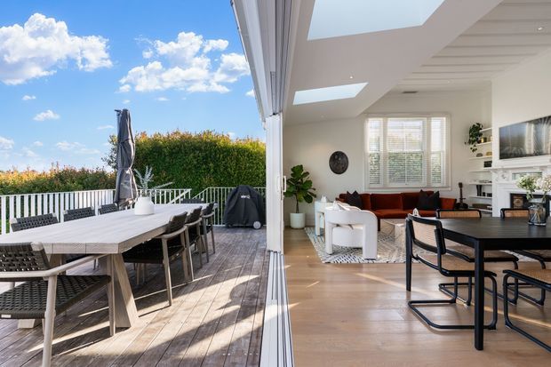Bright contemporary open-plan living and dining area opening onto a timber deck through fully retractable glazed doors. The interior has light timber flooring, white walls and ceiling, skylights, a white fireplace surround, built-in shelving, a dark dining table, and a warm rust-coloured sofa. The deck includes a large outdoor dining setting, with a leafy hedge and blue sky beyond.