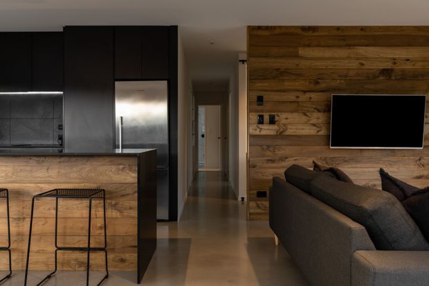 A contemporary open-plan kitchen and living area with dark matte cabinetry, stainless-steel appliances, a timber-clad island and feature wall, black metal bar stools, and a charcoal sectional sofa. A hallway extends beyond the living space, and a wall-mounted television is integrated into the timber feature wall.