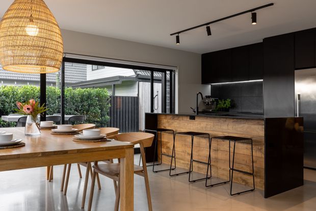 A contemporary open-plan kitchen and dining area with a timber dining table and chairs in the foreground, a timber-fronted island with black metal stools, dark cabinetry, and a dark stone or composite benchtop. Large sliding glass doors open to a planted outdoor area, while a woven pendant light and black track lighting add warmth and contrast.