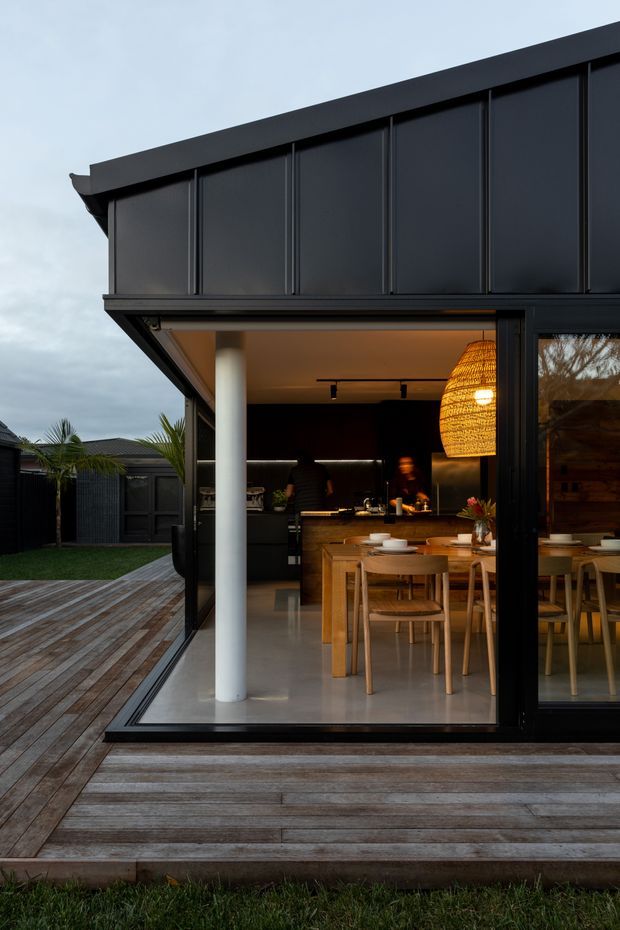 Exterior view of a contemporary dark-clad home opening onto a timber deck. Large black-framed sliding glazing reveals an open-plan dining and kitchen area with a timber dining table, warm woven pendant light, and dark cabinetry. The deck, lawn, fenced garden, and covered outdoor connection are visible, creating a strong indoor-outdoor relationship.