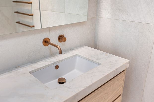 Close-up of a refined bathroom vanity featuring a white integrated rectangular basin set into a pale veined stone benchtop, light timber drawer cabinetry, and wall-mounted copper-toned tapware. Large light grey stone-look wall tiles and a mirror with timber shelving are visible, creating a warm, contemporary finish.