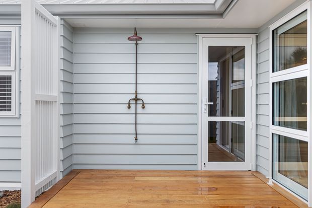Covered exterior patio or entry deck with warm timber decking, pale blue-grey horizontal weatherboard cladding, white-framed glazed door and windows, and a wall-mounted outdoor shower fixture. The space is bright and sheltered, with a clean coastal residential character.