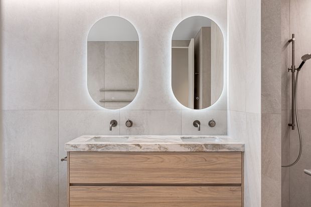Contemporary double-vanity bathroom with pale large-format wall tiles, a timber-look floating vanity, two oval backlit mirrors, wall-mounted brushed metal tapware, and a dark metal handheld shower set.