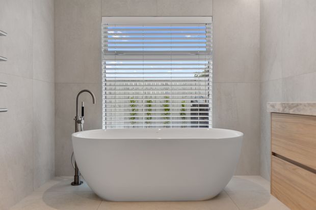 Bright contemporary bathroom featuring a freestanding white bathtub positioned beneath a window with white horizontal blinds, a dark floor-mounted bath filler, pale large-format wall and floor tiles, and a light timber floating vanity with a stone benchtop at right.