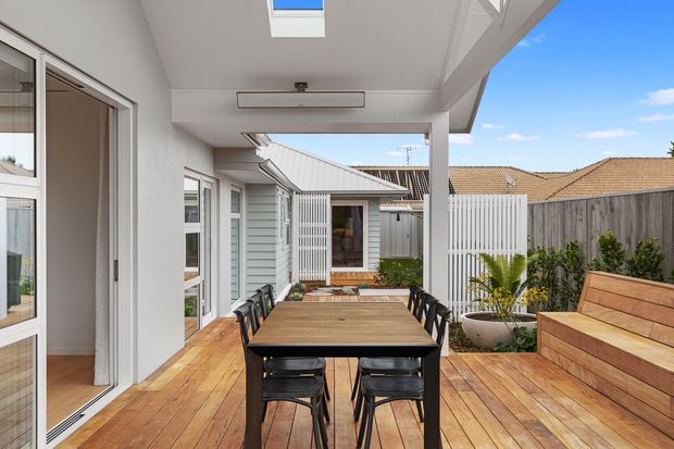 A bright covered outdoor dining terrace with a large timber dining table and black chairs on a warm timber deck. The space is enclosed by light-painted weatherboard walls, white-framed glazing, privacy fencing, and a built-in timber bench with planters. A roof-mounted skylight and overhead structure provide shelter, while neighbouring residential rooflines are visible beyond.