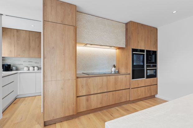 Bright contemporary kitchen with a full-height wall of warm timber cabinetry, integrated black oven and microwave, concealed rangehood, pale textured splashback, and a compact stone benchtop. A white island is visible in the foreground, with a secondary galley kitchen area seen through the opening at left. Light timber flooring and white walls create a clean, warm, minimalist interior.