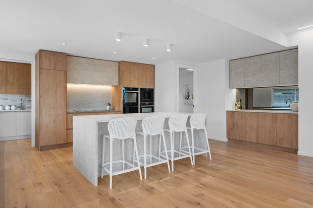 Bright contemporary kitchen with a large pale stone island and four white bar stools, warm timber flooring, and extensive timber cabinetry. The rear wall includes integrated black appliances, a softly illuminated textured splashback, and additional cabinetry with a window above the benchtop. Recessed and small surface-mounted ceiling lights create an uncluttered, airy interior.