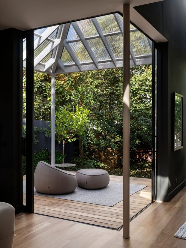 A modern indoor-outdoor lounge opening onto a timber deck and lush enclosed garden. Full-height black-framed glazing and a glazed sloping roof create a bright connection to the greenery, while a sculptural curved lounge chair, matching ottoman, small side table and neutral rug furnish the deck. The interior has dark walls, warm timber flooring and a slender structural post.