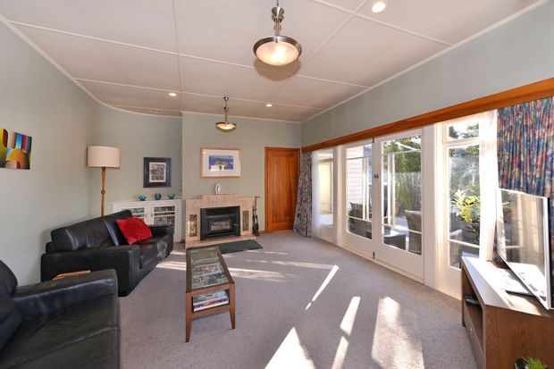 A bright traditional living room with pale grey walls, carpeted flooring, a decorative tiled fireplace, black leather sofas, a wooden coffee table, and a timber media console. Full-height glazed doors and windows open to a covered outdoor area, while patterned curtains and timber trim add warmth. The room has a low tiled ceiling with recessed lighting and pendant fixtures.