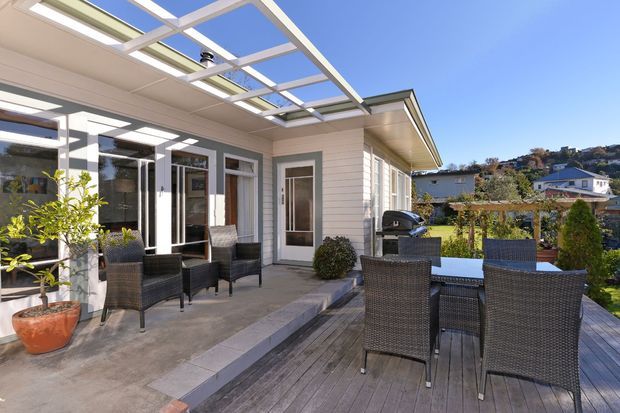 A bright outdoor entertaining area adjoining a house, with a timber deck furnished with a dark woven dining table and chairs. A covered patio with a translucent roof structure, outdoor seating, potted plants, barbecue, glazed doors, and pale weatherboard cladding overlooks a suburban hillside neighbourhood.