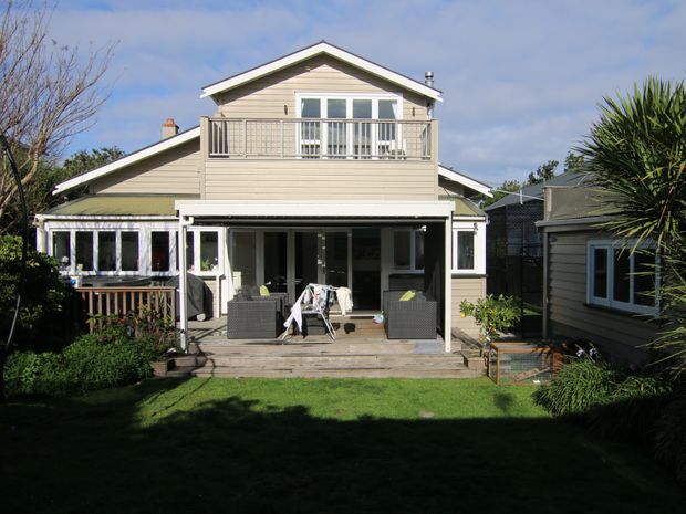 Rear exterior view of a light-coloured, multi-level weatherboard house with a prominent upper gable, enclosed balcony, covered ground-level veranda, timber deck, and surrounding garden. The image is photographed from the backyard and shows a small detached weatherboard structure to the right.