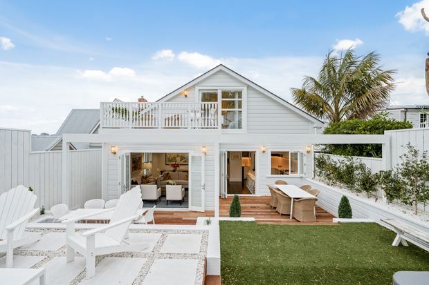 Exterior view of a bright white two-storey coastal-style house with horizontal weatherboard cladding, a prominent gabled roof, upper balcony, large glazed openings, and a landscaped outdoor entertaining area. The garden includes a tiled patio with white Adirondack chairs, timber decking with outdoor dining furniture, lawn, white fencing, and a palm tree.