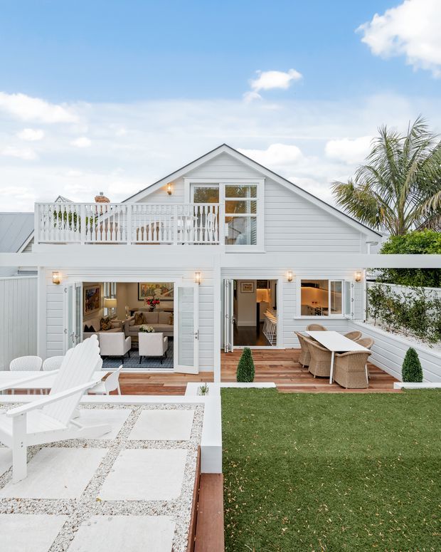 A bright exterior view of a white two-storey coastal-style home with horizontal weatherboard cladding and a prominent gabled roof. The property features a large upper balcony with white balustrading, multiple glazed doors opening onto timber decks, outdoor dining furniture, a paved patio with white loungers, and a manicured lawn. Interior living and dining areas are visible through the open doors.
