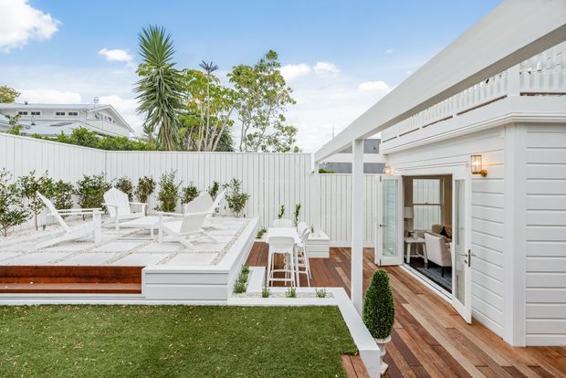 Bright rear courtyard and terrace of a contemporary white-clad home, featuring a raised outdoor entertaining platform with white lounge chairs, a small dining setting, timber decking, manicured lawn, clipped topiary, and an open glazed doorway into the interior. The setting has a clean resort-like aesthetic with strong white and timber finishes under a clear blue sky.