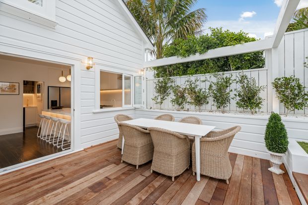 A bright outdoor dining terrace beside a white weatherboard house. The timber deck is furnished with a white rectangular dining table and woven wicker chairs, enclosed by a white fence and trellis-style planting wall. White pergola beams frame the space, with a kitchen and living area visible through the open glass doors. Palm foliage and clear blue sky contribute to a relaxed coastal atmosphere.