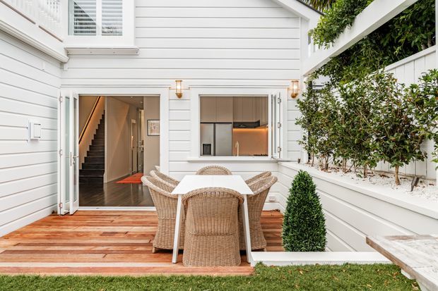 A bright, enclosed courtyard-style outdoor dining area beside a white weatherboard house. The space features a timber deck, white rendered or painted raised planter walls, climbing greenery, a white outdoor dining table with woven wicker chairs, and a compact clipped shrub. Open doors connect the courtyard to an interior hallway and kitchen, while a second-storey window and stairway are visible.