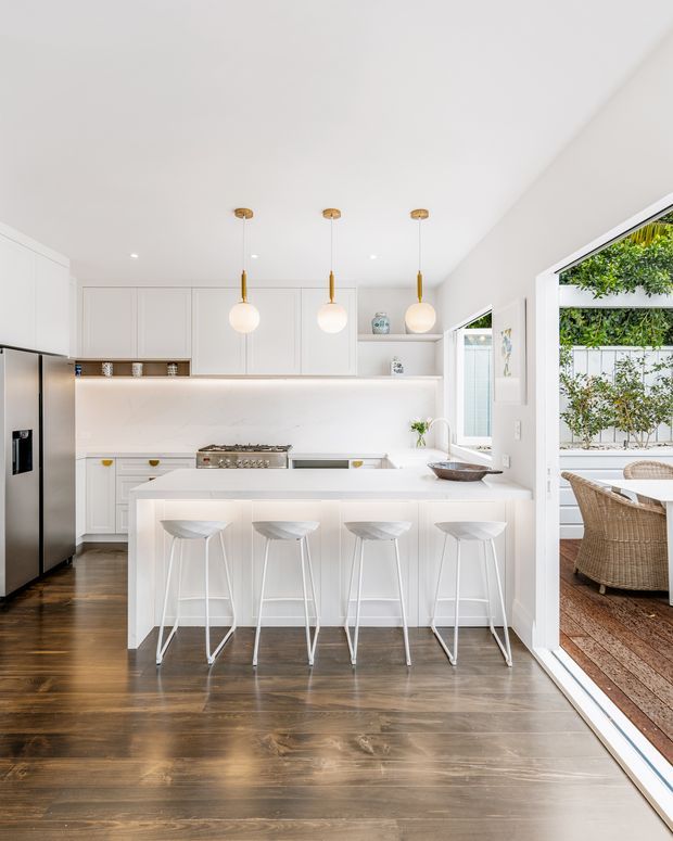 Bright contemporary white kitchen with a large island and four white stools, white paneled cabinetry, integrated shelving, stainless-steel refrigerator and range, and dark timber flooring. Three globe pendant lights hang above the island. Full-height glazed doors open to a furnished outdoor deck or patio, bringing in abundant natural light and greenery.