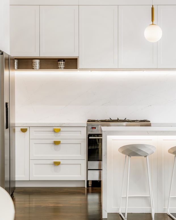 A bright, compact contemporary kitchen with white framed cabinetry, a large white marble-look splashback and island, integrated under-cabinet lighting, stainless-steel cooking appliances, and a dark timber floor. The island includes white decorative paneling and seating for two with sculptural grey stools. A small open timber display shelf and a brass globe pendant add warm contrast.