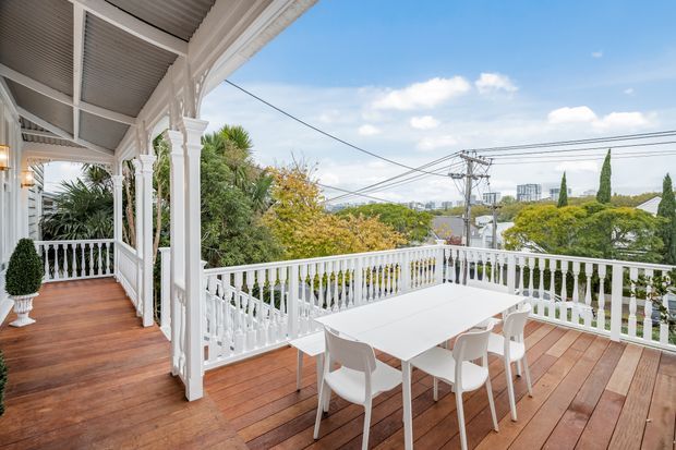 An elevated outdoor timber deck and covered veranda with ornate white painted timber columns, balustrades, and louvred ceiling panels. A white outdoor dining table and chairs occupy the deck, which overlooks leafy suburban surroundings and a distant urban skyline.