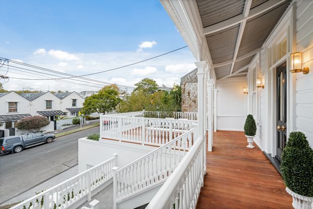 A bright covered verandah or balcony running along the front of a white multi-storey residence. The space features warm timber decking, ornate white balustrades and columns, white weatherboard walls, decorative exterior lighting, and potted topiary. Stairs connect to a lower terrace, with neighbouring pitched-roof homes and a residential street visible beyond.