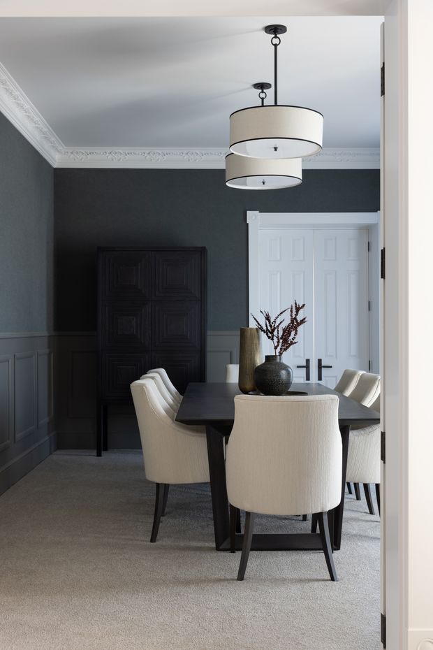 A refined formal dining room viewed through a doorway, featuring a dark rectangular dining table surrounded by upholstered cream dining chairs. The room has charcoal-grey walls with white traditional wall panelling and ornate crown moulding, a grey carpet, a dark decorative cabinet, white paneled double doors, and paired cream drum pendant lights. A central vase arrangement provides a subtle natural accent.