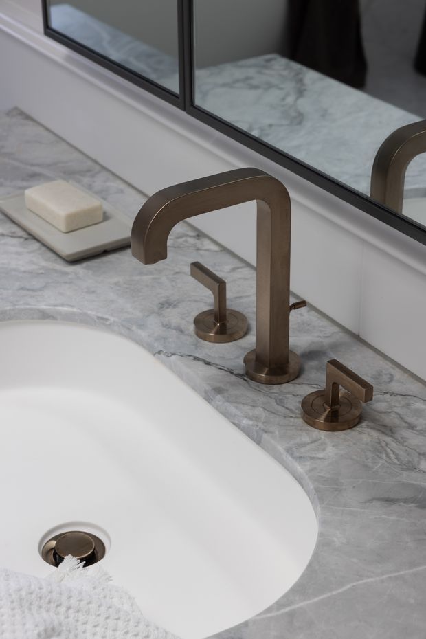 Close-up of a refined bathroom vanity featuring a white integrated or inset basin set into a grey-white veined marble countertop. A sculptural brushed bronze or dark brass wall-mounted style mixer tap and separate controls sit beside the basin, with a large black-framed mirror above and a white soap dish on the counter.