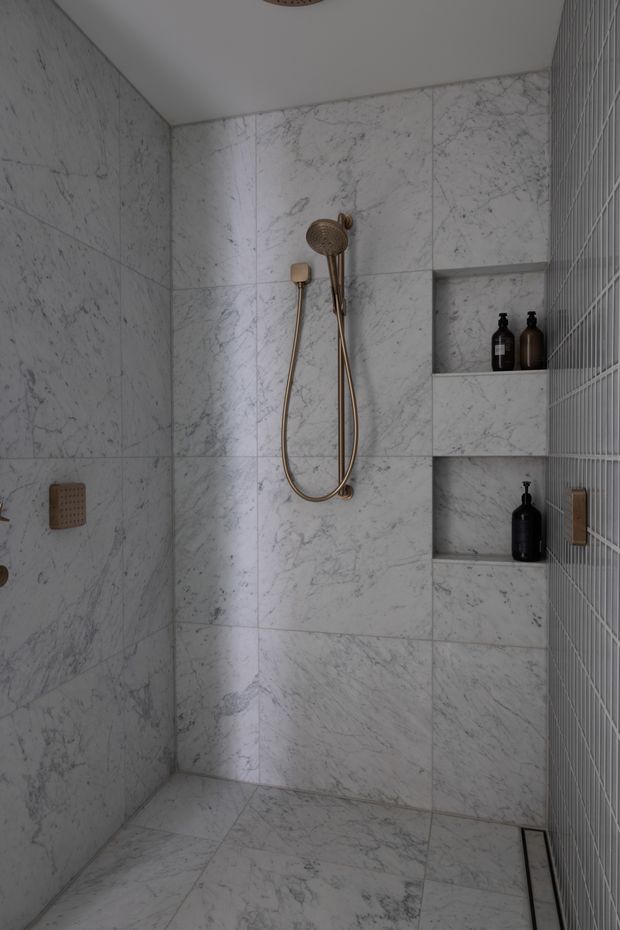 A compact contemporary walk-in shower lined with white-grey veined marble-look tiles on the walls and floor. The shower features brushed brass fixtures, recessed tiled niches with dark bottles, a linear floor drain, and a narrow vertical tile mosaic on the right wall.