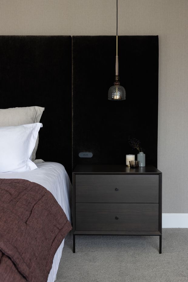 A close-up view of a refined bedroom corner featuring a bed with white bedding and a dark brown textured throw, a tall black upholstered headboard, and a dark wood two-drawer bedside cabinet. A small pendant light hangs above the nightstand, with simple decorative vessels and a small plant on top. The room has neutral textured walls, white skirting boards, and grey carpet, creating a calm, understated atmosphere.