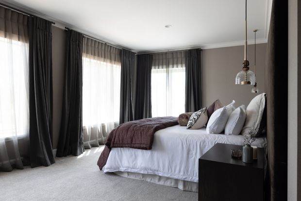A refined bedroom with a large upholstered bed dressed in white bedding and layered grey-purple throws and cushions. Floor-to-ceiling windows are framed by full dark charcoal curtains and sheer drapes, while a dark bedside table and pendant light complement the taupe-grey walls and pale carpet. The room is softly lit and elegantly styled.