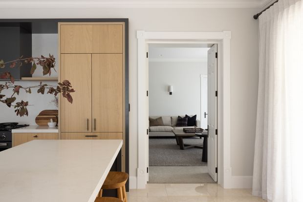 Bright, refined open-plan kitchen interior with a large pale stone island, integrated timber cabinetry framed in dark trim, and a view through a decorative white-trimmed doorway into a calm living room. Soft sheer curtains, neutral finishes, and natural timber accents create a contemporary transitional feel.