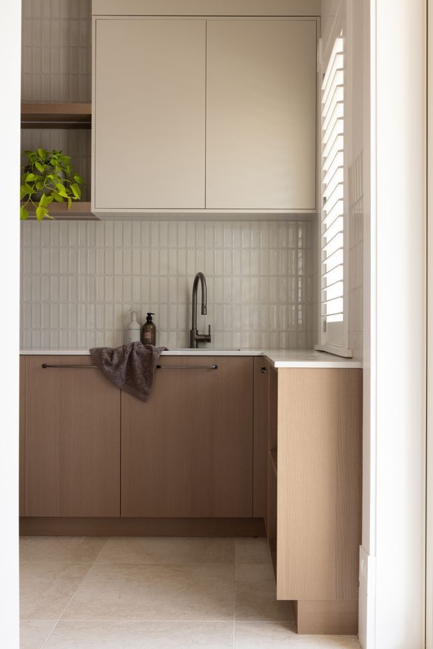 Compact, softly lit contemporary kitchen or kitchenette with warm wood-grain lower cabinetry, pale cream upper cabinets, a light stone benchtop, and a vertically ridged pale tile splashback. A dark gooseneck tap and undermount sink sit beneath a louvered window, with open shelving and a small plant at left. Large beige floor tiles and restrained detailing create a calm, warm interior.
